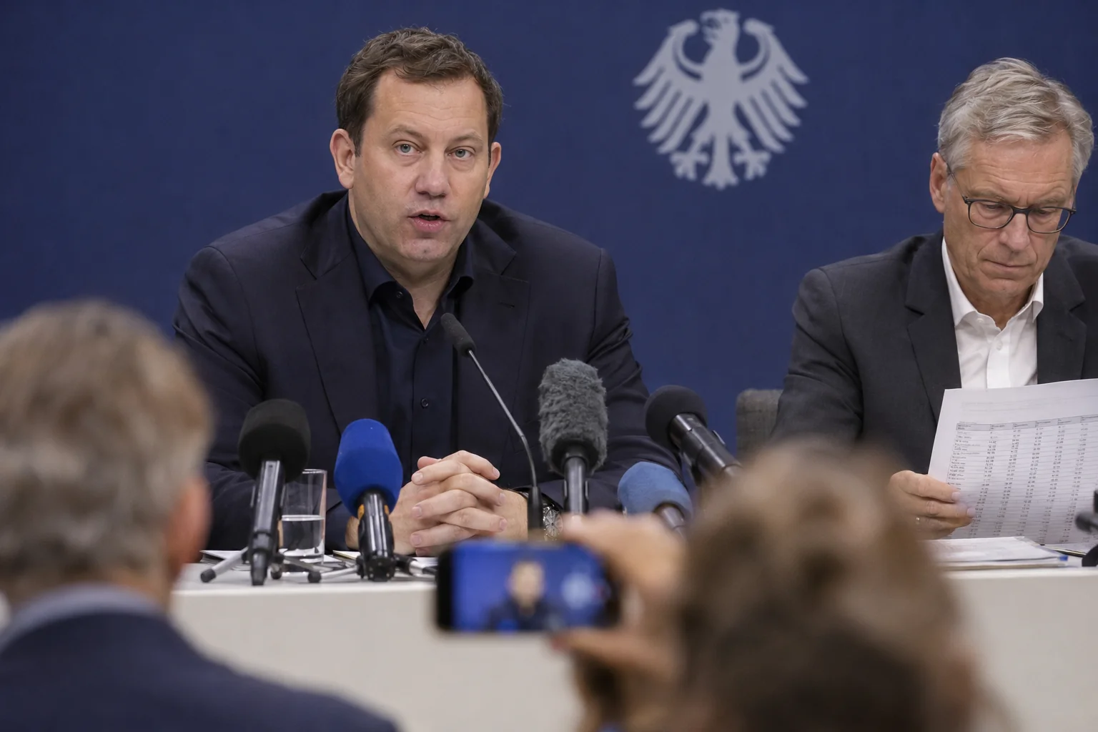 KI-generierte Illustration: Finanzminister Lars Klingbeil spricht bei einer Pressekonferenz vor dem Bundesadler in Mikrofone, neben ihm ein Mann mit Zahlentabelle