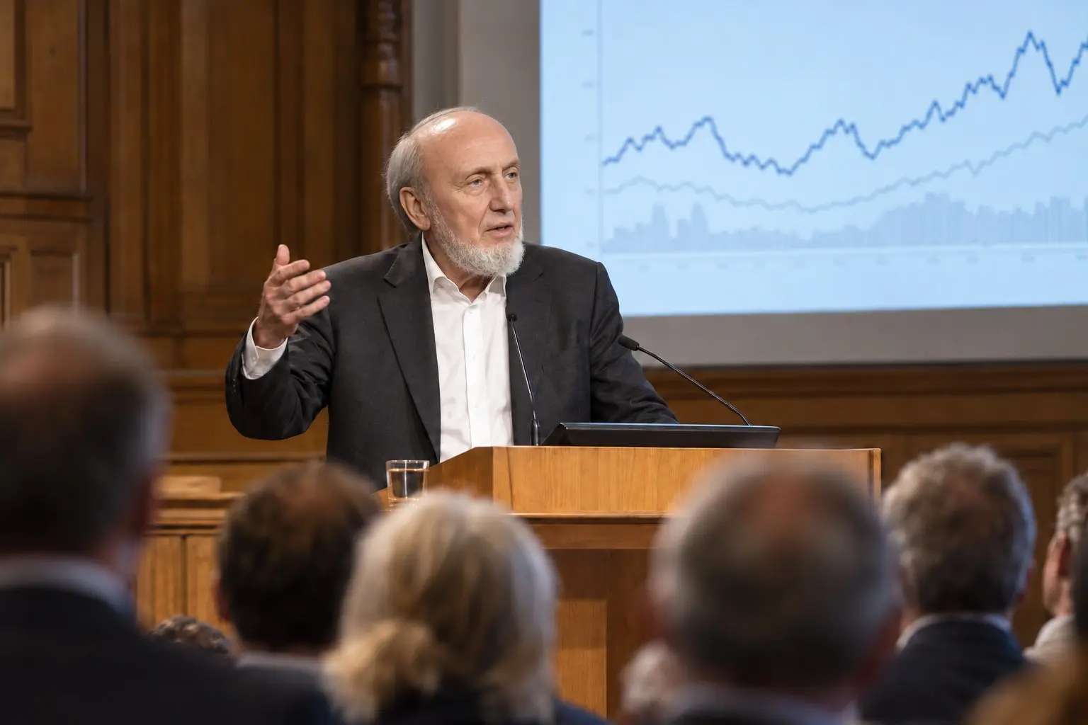 KI-generierte Illustration: Hans-Werner Sinn am Rednerpult eines holzvertäfelten Hörsaals, eine Hand erhoben, dahinter eine Leinwand mit Kurvendiagramm, Zuhörer unscharf im Vordergrund; Nachrichtenfoto-Stil.
